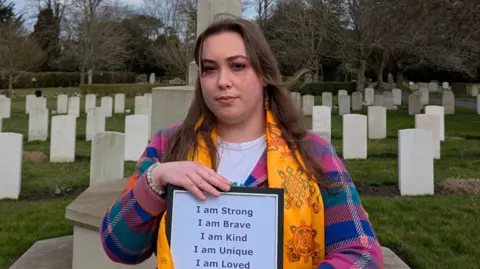 Ed Gold/Positive Futures Claire wearing a colourful chequered jumper and a yellow scarf while standing in front of graves in a graveyard. She has long brown hair and is holding a piece of paper that says she is strong, brave, kind, unique and loved.